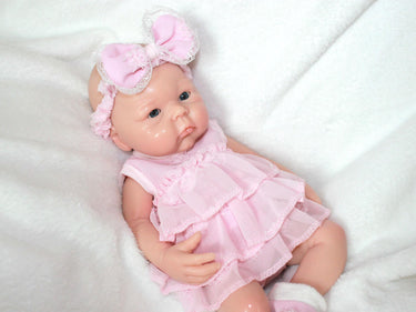 A baby doll dressed in a pink dress and matching headband, sitting against a soft background.